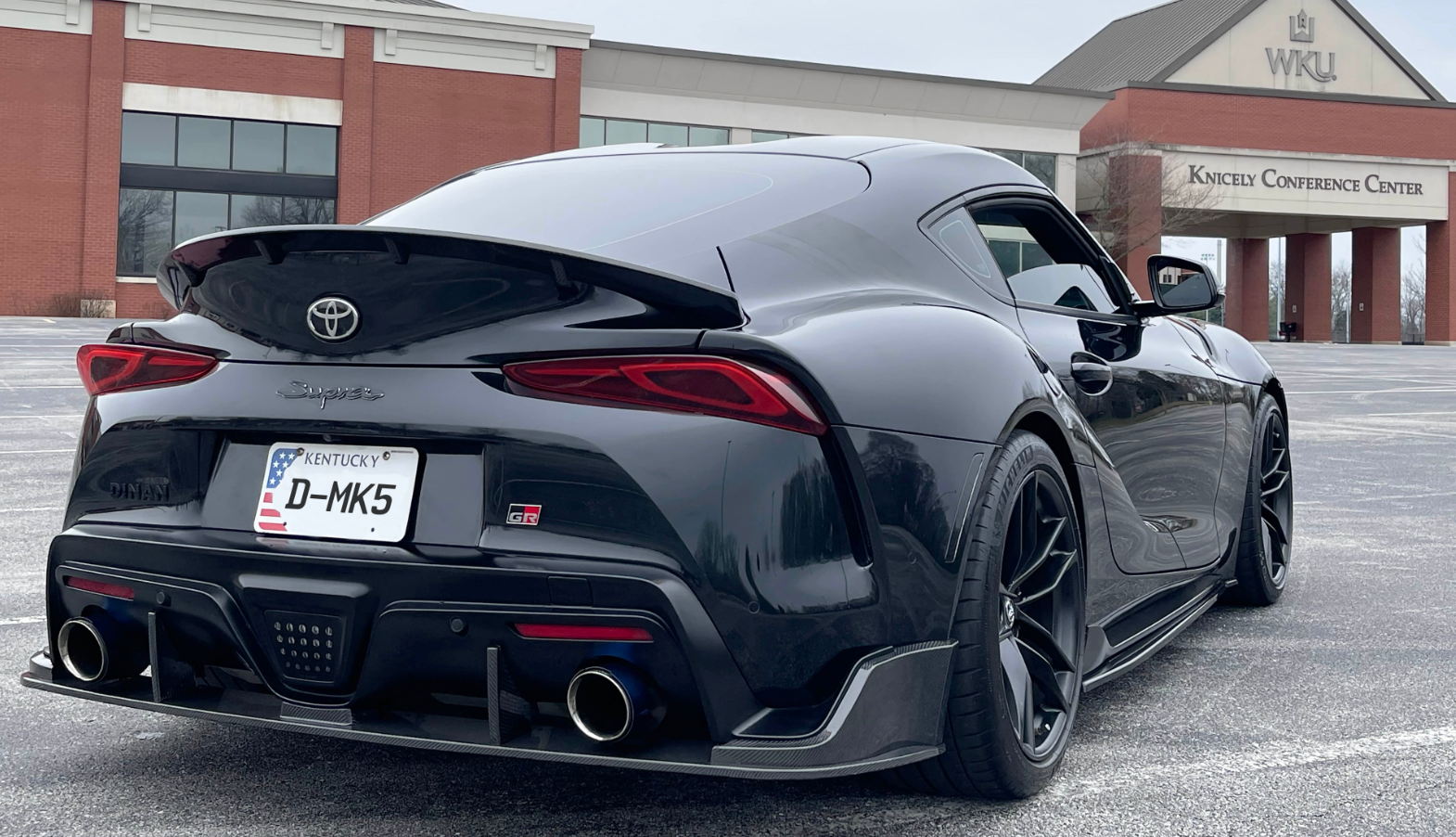 A back view of a black car fitted with Dinan Rear Deck Spoiler for Toyota GR Supra 2020-2025 on the road in front of Knicely Conference Center