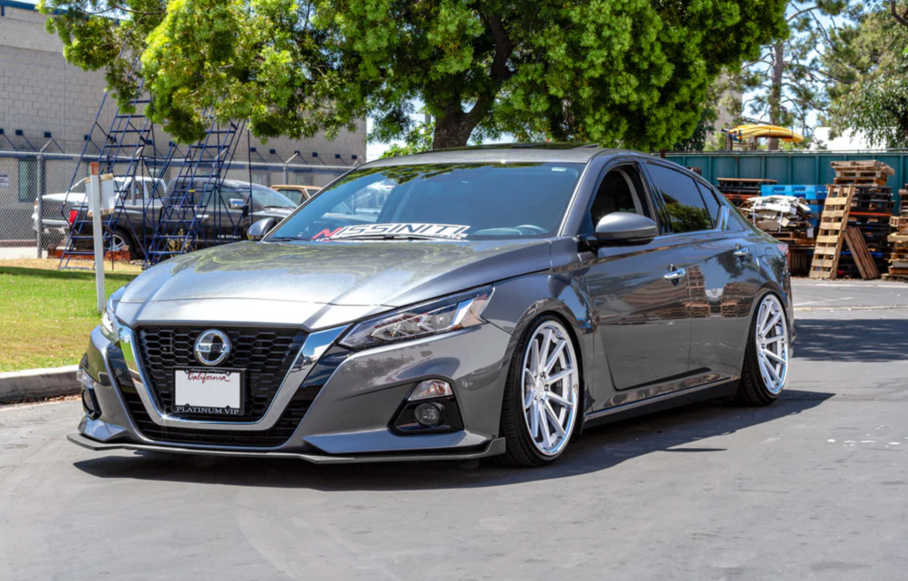 A front wide view of a silver car fitted with Stillen Front Splitter [Matte Black] for Nissan Altima 2019-2022 on the ground outside with a big tree behind it