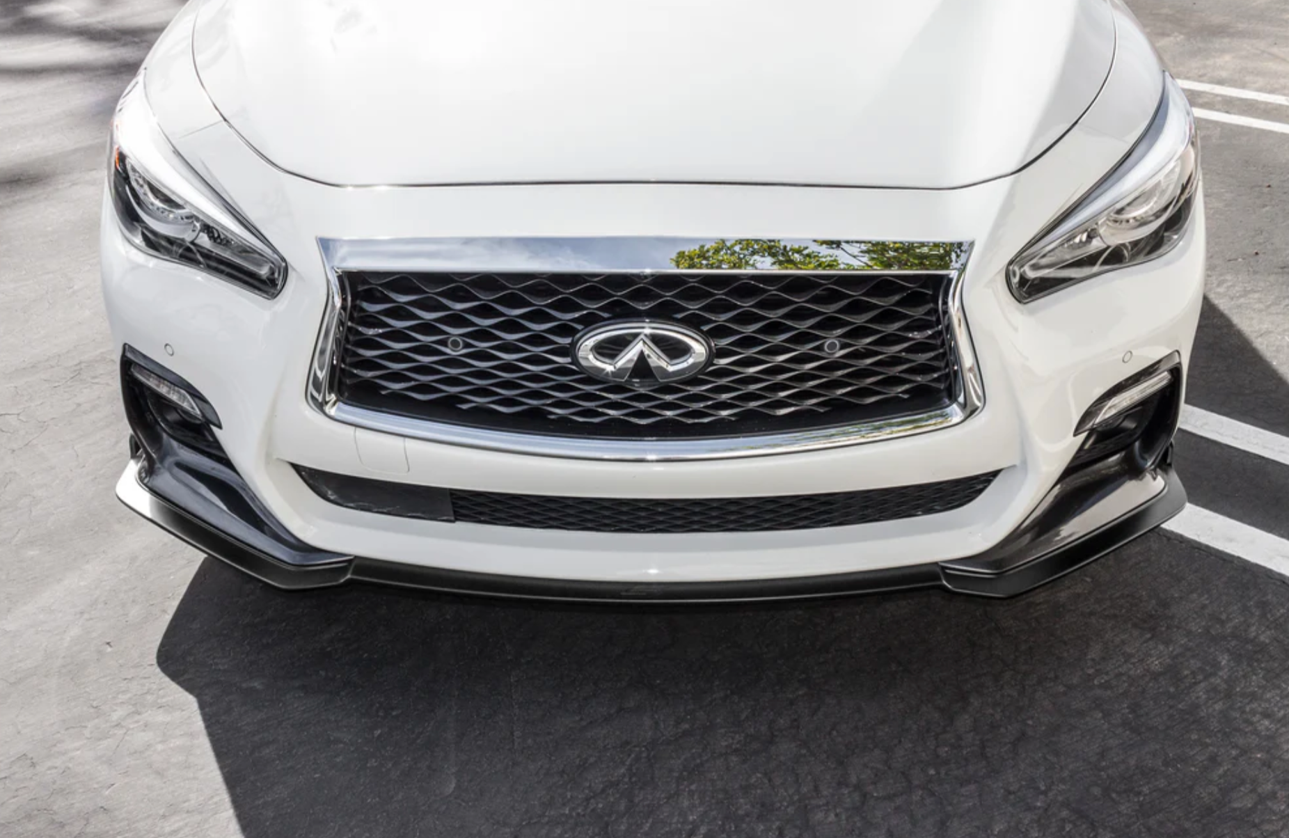 A close up view of the front of a white car fitted with Stillen Front Splitter [Matte Black] for Infiniti Q50 2018-2024 on the ground