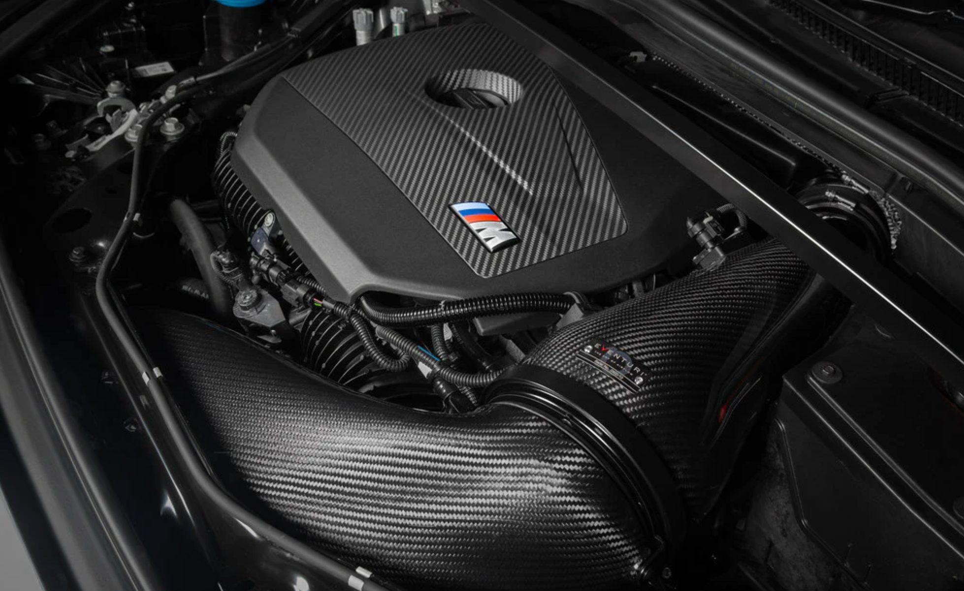 A top side view of an open bonnet of a car fitted with Eventuri Black Carbon Intake System for BMW F7X M135i M235i / U1X X1 X2 M35i & MINI F6X S JCW 2020-2025 on the ground