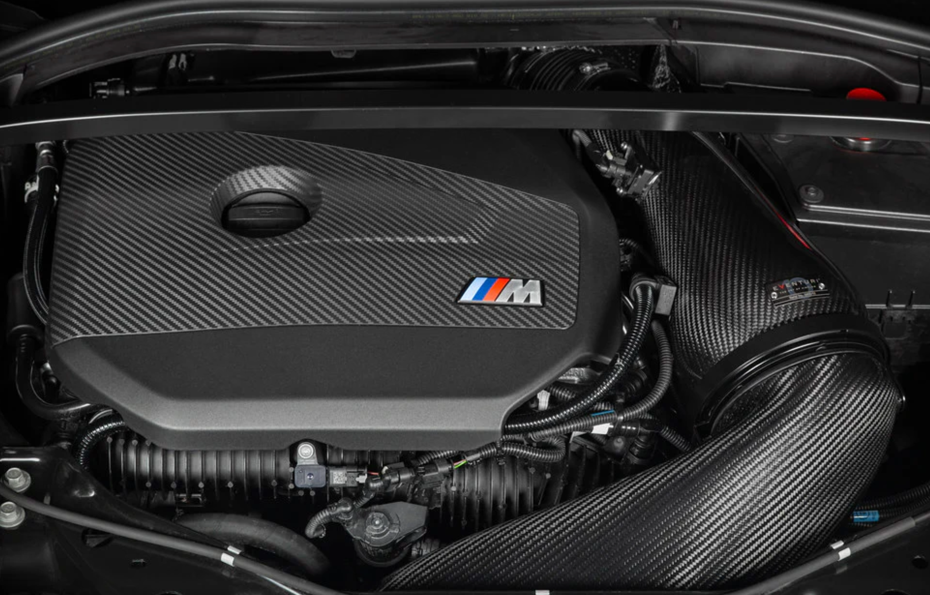 A close up view of an open bonnet of a car fitted with Eventuri Black Carbon Intake System for BMW F7X M135i M235i / U1X X1 X2 M35i & MINI F6X S JCW 2020-2025 on the ground