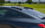 A side close up view of a black car fitted with AutoTecknic Carbon Fiber Roof Fin Set - BMW G82 M4 2021-2025 on the ground with greenery behind