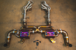 A top-down view of a complete automotive exhaust system on a concrete floor, including headers, an X-pipe, two iridescent purple and gold resonators, and curved tailpipes, with an electronic valve controller kit positioned in the center.