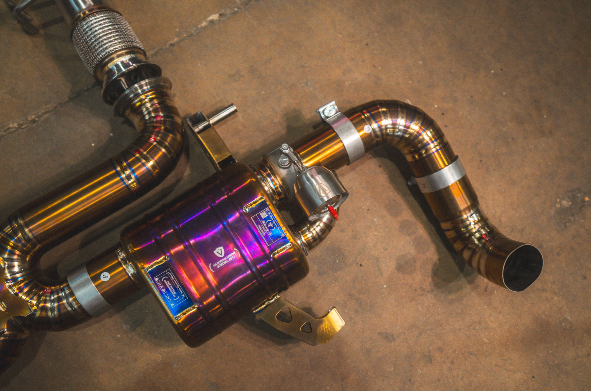 A detailed close-up of a heat-treated, iridescent purple and gold exhaust muffler featuring a vacuum-actuated valve, a product badge, and sectional welded piping.