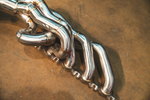 A side-angle view of a polished metal exhaust header featuring multiple curved primary tubes and a thick engine mounting flange.