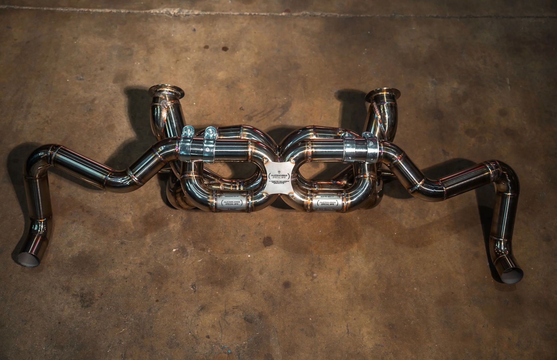 A top-down view of a polished stainless steel exhaust system featuring a complex central X-pipe crossover with four internal pipe loops and dual out-swept tailpipes on a concrete floor.
