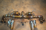 An overhead shot of a titanium exhaust system with a heat-colored golden finish, featuring a central X-pipe, dual mufflers, four large separate exhaust tips, and a handheld remote control with a wiring harness.
