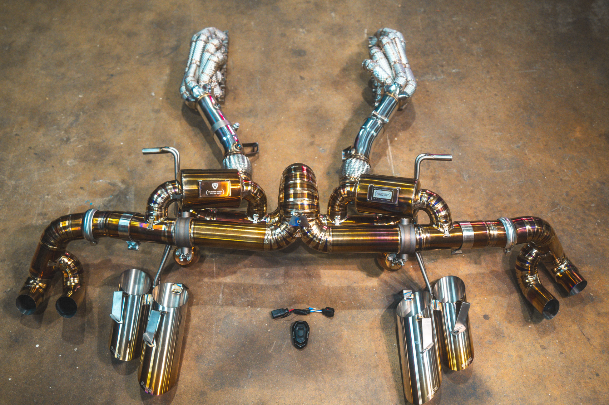 A full assembly view of a titanium exhaust system laid out on a concrete floor, featuring heat-shielded headers connected to an X-pipe, dual mufflers, and four large exhaust tips, with a controller kit in the foreground.