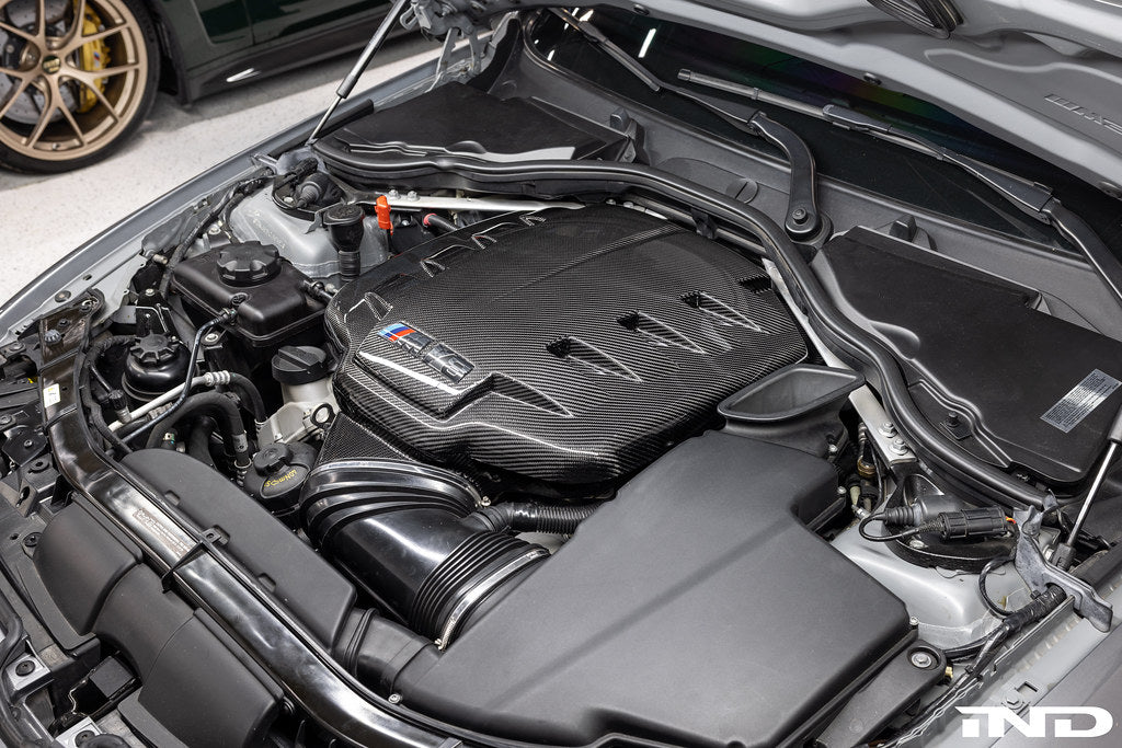 Black carbon fiber inlet plenum with curved design installed in BMW M3 engine bay with visible intake tubes - Eventuri inlet plenum for BMW M3