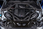 Carbon fiber strut brace installed in engine bay spanning across shock towers with diagonal bracing pattern - Eventuri strut brace for BMW M2