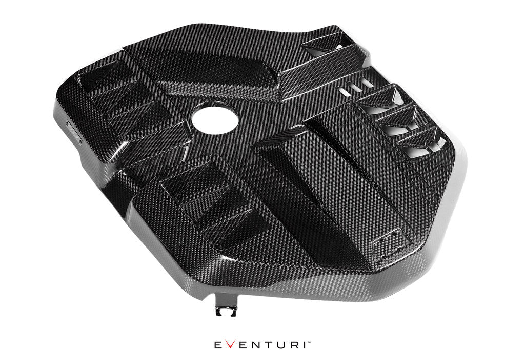 Black carbon fiber engine cover with geometric sculpted design, central circular opening, and integrated ventilation louvers on clean white background - Eventuri engine cover for BMW M2
