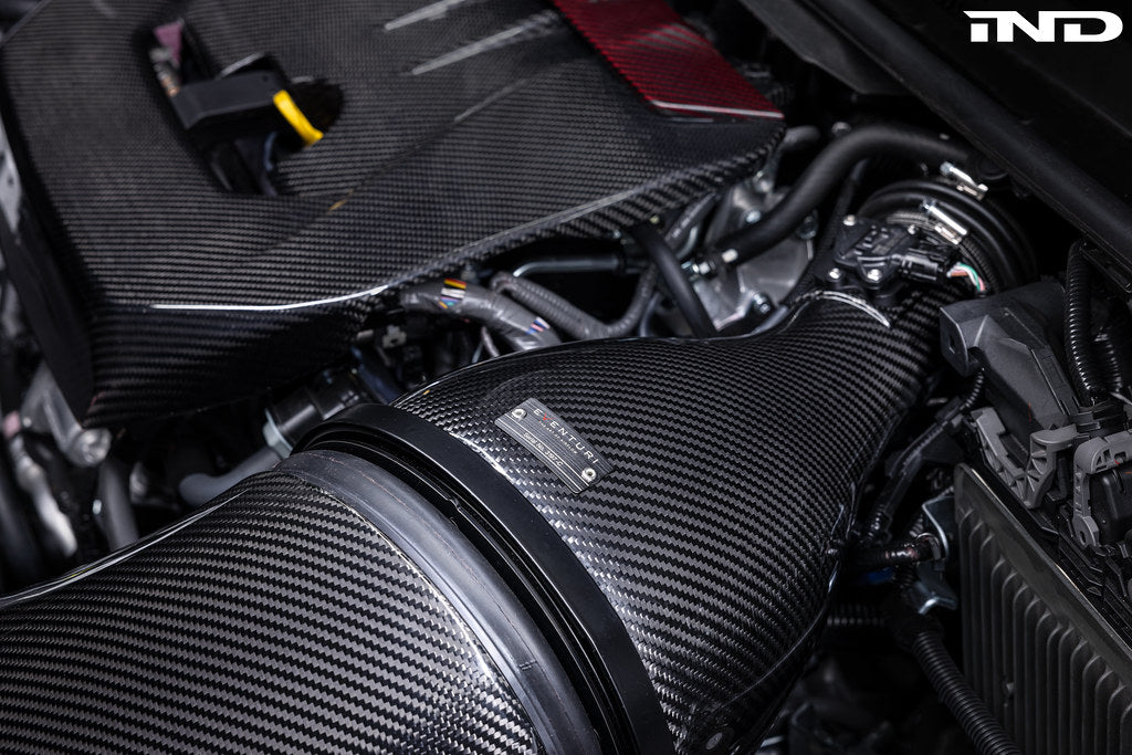 Carbon fiber intake system with cylindrical air filter installed in GR Corolla engine bay showing detailed weave pattern - Eventuri carbon intake for Toyota GR Corolla