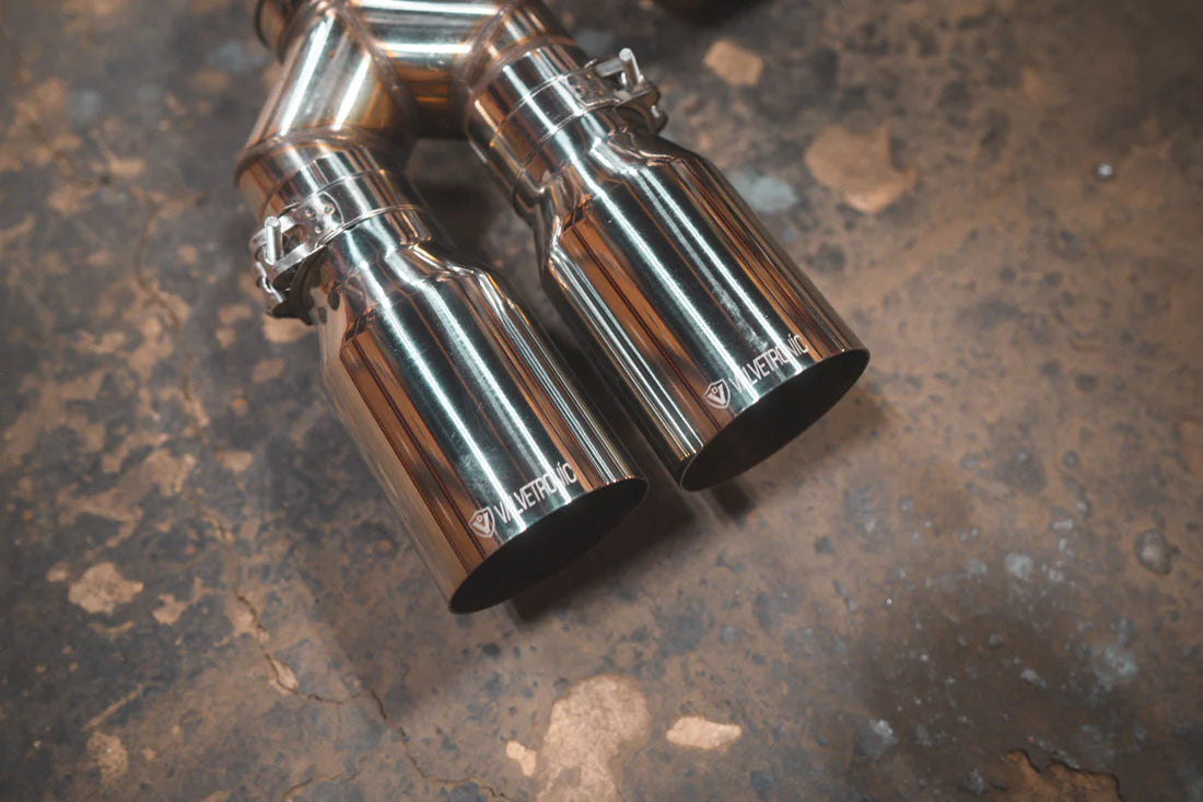 A close up view of Valvetronic Designs Equal Length Valved Sport Exhaust System's Chrome exhaust tips on the ground (for BMW M3 F80, M4 F82, M4 F83).