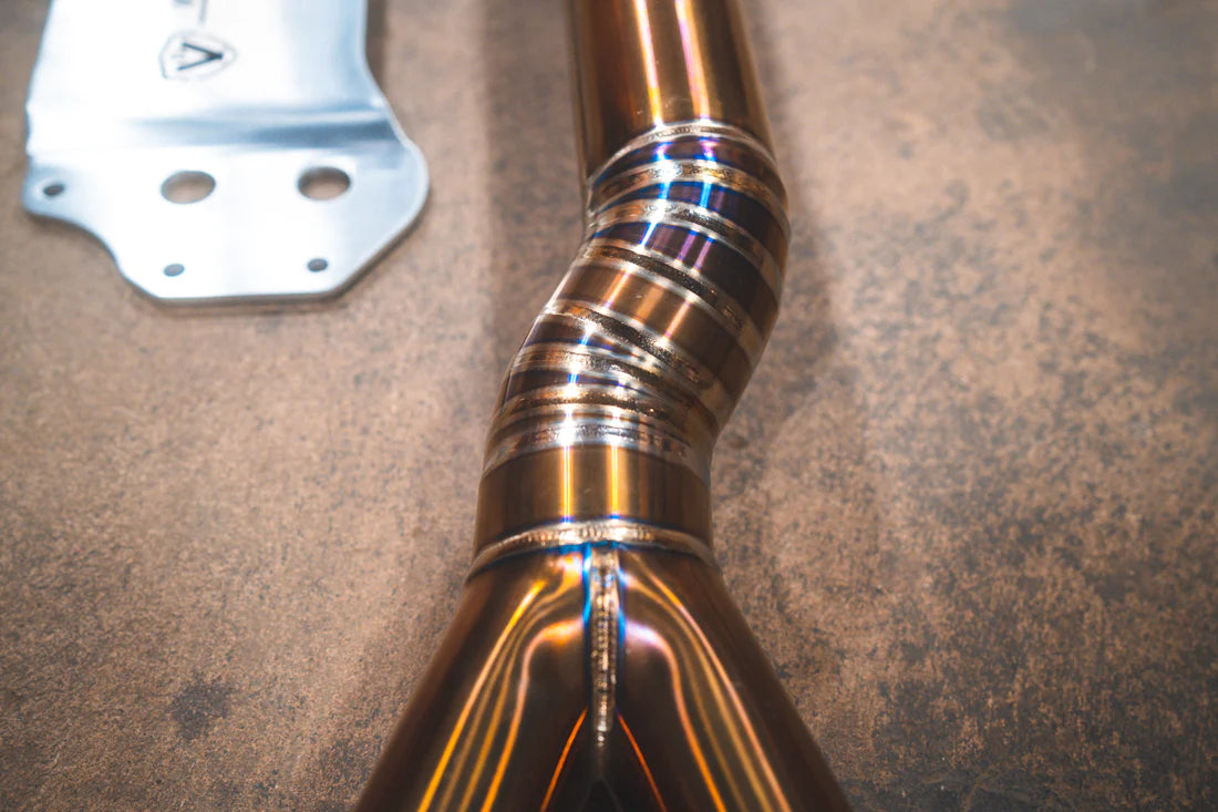 A close up view of Valvetronic Designs Equal Length Midpipe in Grade 5 Titanium on the ground (for BMW G8x M3 / M4).