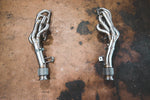 A close-up of two Valvetronic Designs Free Flow Headers (for Audi R8 V10 / Lamborghini Gallardo LP / Lamborghini Huracan) exhaust pipes facing each other on the ground.	