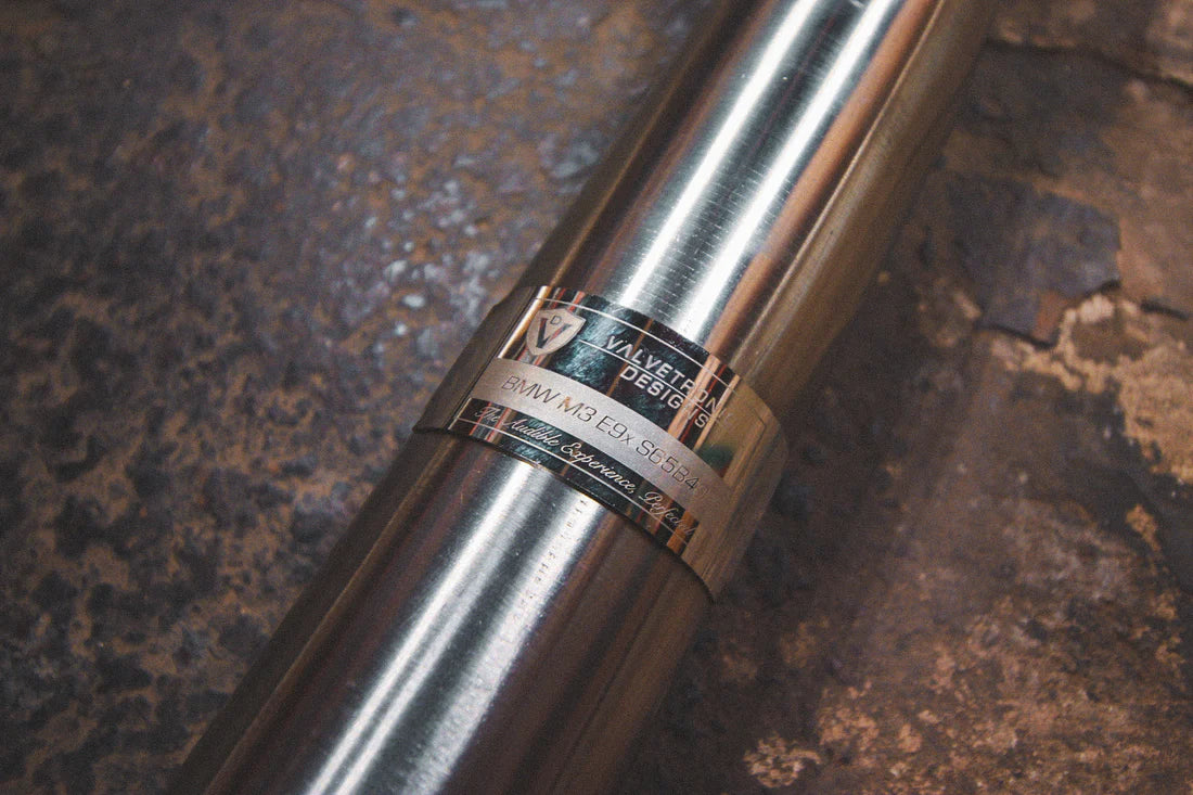 A close up view of Valvetronic Designs Midpipe V2, showing the brand's logo, on the ground (for BMW M3 E9x).