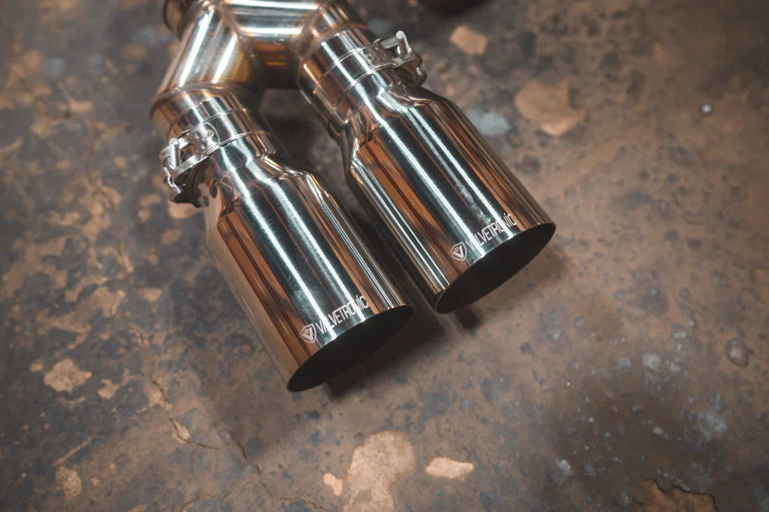 A close up view of Valvetronic Designs Equal Length Valved Sport Exhaust System's Chrome exhaust tips on the ground (for BMW M3 F80, M4 F82, M4 F83).