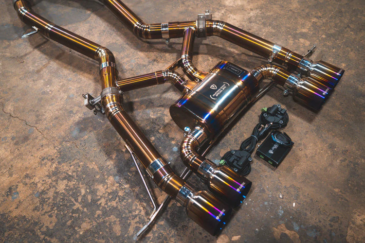 A close view of Valvetronic Designs Axleback GRADE 5 TITANIUM Valved Sport Exhaust System with Raw Titanium Burnt exhaust tips on the ground (for BMW G8x M3 / M4).
