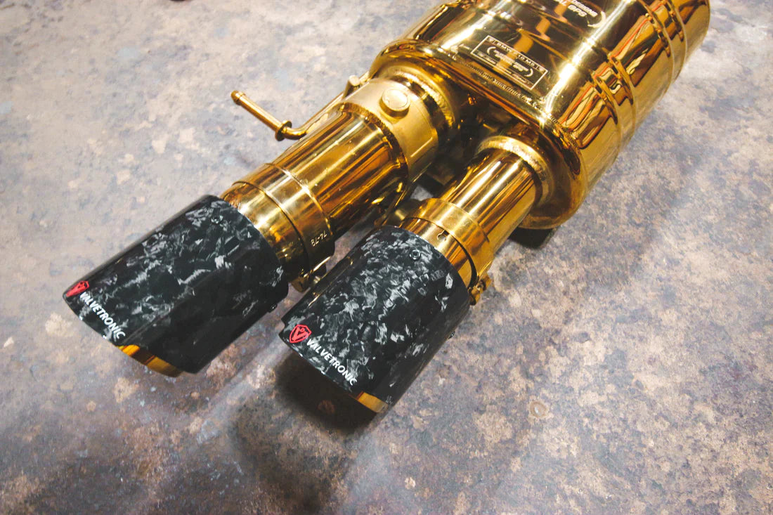 A close overview of Valvetronic Designs Anodized Gold Valved Sport Exhaust System's muffler and Forged Carbon Fiber exhaust tips on the ground (for BMW F10 M5 / F12 M6 / F06 M6).