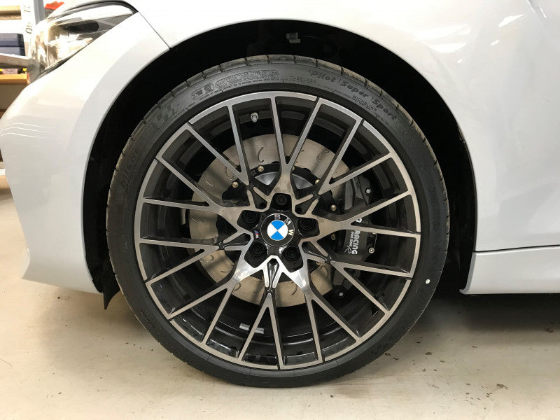 A close up view of a white BMW car with the front wheel fitted with AP Racing by Essex Radi-CAL ENP Competition Brake Kit (Front CP9660/372mm)- BMW F87 M2 & M2 Competition, F80 M3, F82 M4