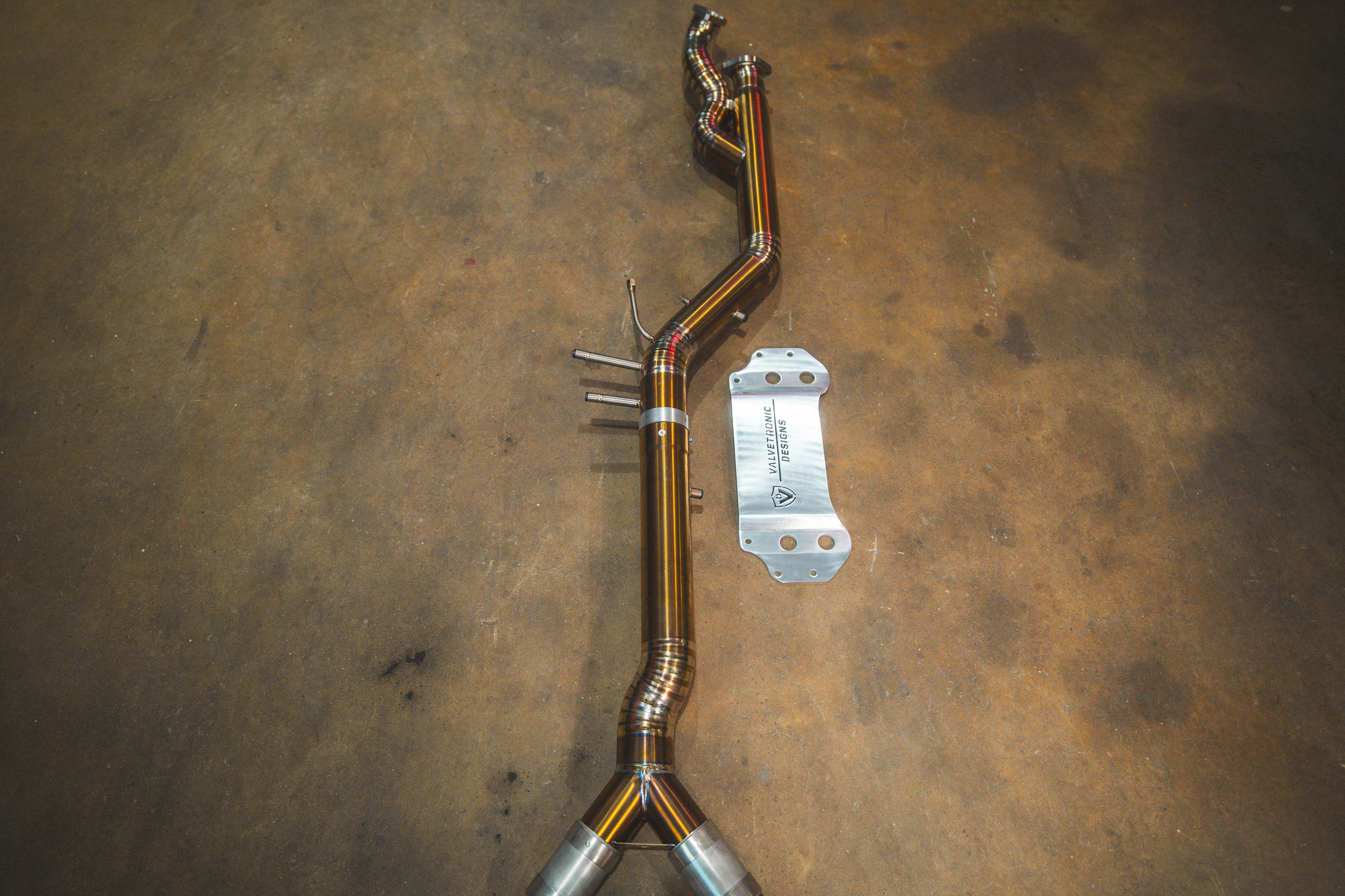 A titanium Valvetronic Designs exhaust mid pipe for a BMW G87 M2, showing distinctive heat-tinted welds and a bronze-hued finish, laid out on a concrete floor with a matching silver heat shield.